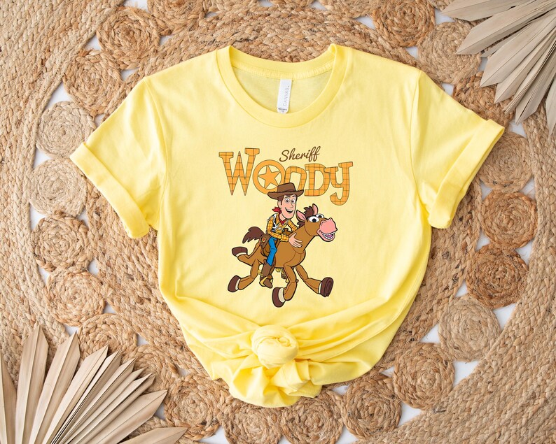 May include: A yellow t-shirt featuring a graphic of Sheriff Woody riding a horse. The text "Sheriff Woody" is above the illustration. The shirt is made of a soft material and has rolled-up sleeves.
