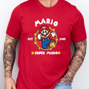 May include: Red t-shirt featuring a graphic of Mario from Super Mario Bros. The design includes the word "MARIO" in white, the year "1985", and the phrase "SUPER MARIO". The character is in his classic pose.