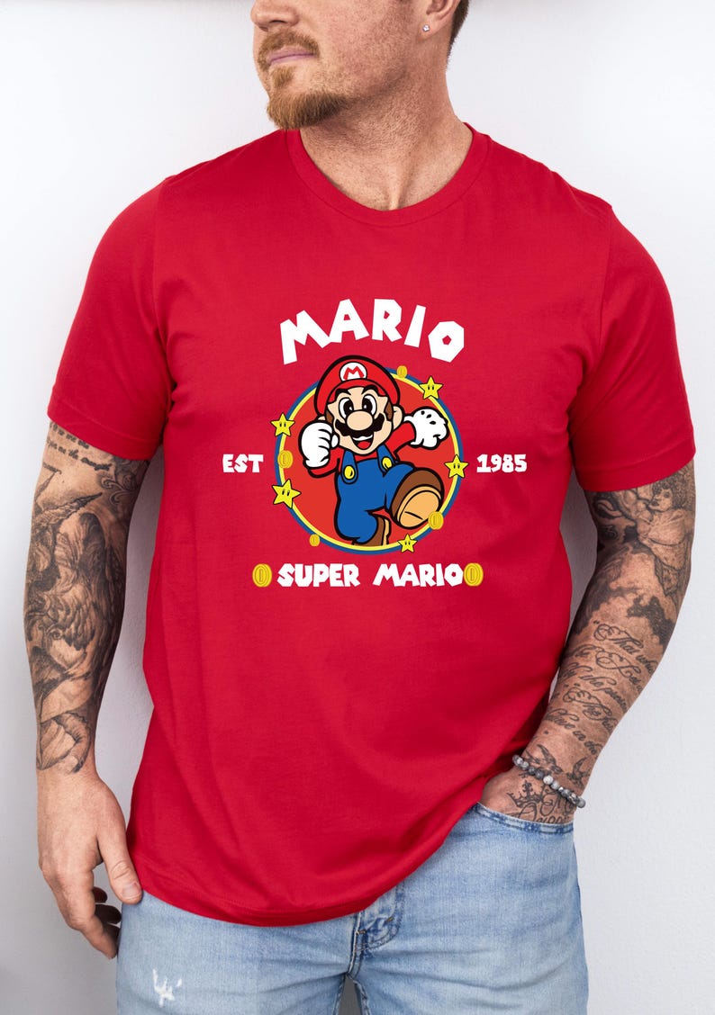 May include: Red t-shirt featuring a graphic of Mario from Super Mario Bros. The design includes the word "MARIO" in white, the year "1985", and the phrase "SUPER MARIO". The character is in his classic pose.