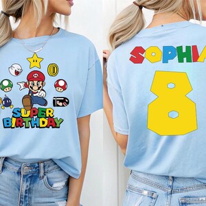K&ouml;nnte beinhalten: Hellblaues T-Shirt mit einer Grafik eines Videospielcharakters und dem Schriftzug "SUPER BIRTHDAY". Auf der R&uuml;ckseite des Shirts stehen der Name "SOPHIA" und die Zahl "8" in Gelb.