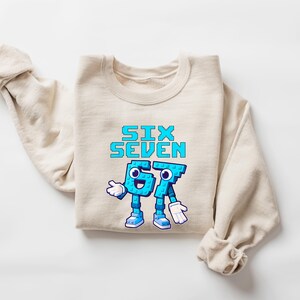May include: Beige crewneck sweatshirt with the text "SIX SEVEN" in pixelated blue letters. The numbers "6" and "7" are personified with arms, legs, and faces. The sweatshirt is folded on a white surface.