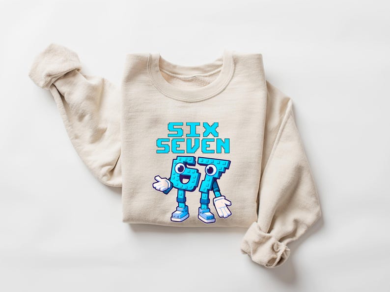May include: Beige crewneck sweatshirt with the text "SIX SEVEN" in pixelated blue letters. The numbers "6" and "7" are personified with arms, legs, and faces. The sweatshirt is folded on a white surface.