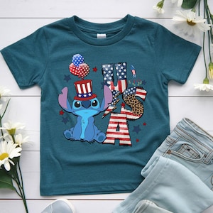 K&ouml;nnte beinhalten: T&uuml;rkisfarbenes T-Shirt mit einer Zeichentrickfigur, die einen patriotischen Hut tr&auml;gt und Luftballons h&auml;lt. Das Wort "USA" ist mit Mustern der amerikanischen Flagge und Leopardenmuster gestaltet. Rote und blaue Sterne und Feuerwerk vervollst&auml;ndigen das Design.
