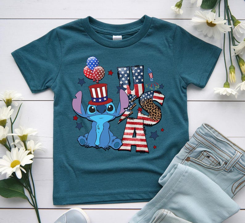 K&ouml;nnte beinhalten: T&uuml;rkisfarbenes T-Shirt mit einer Zeichentrickfigur, die einen patriotischen Hut tr&auml;gt und Luftballons h&auml;lt. Das Wort "USA" ist mit Mustern der amerikanischen Flagge und Leopardenmuster gestaltet. Rote und blaue Sterne und Feuerwerk vervollst&auml;ndigen das Design.