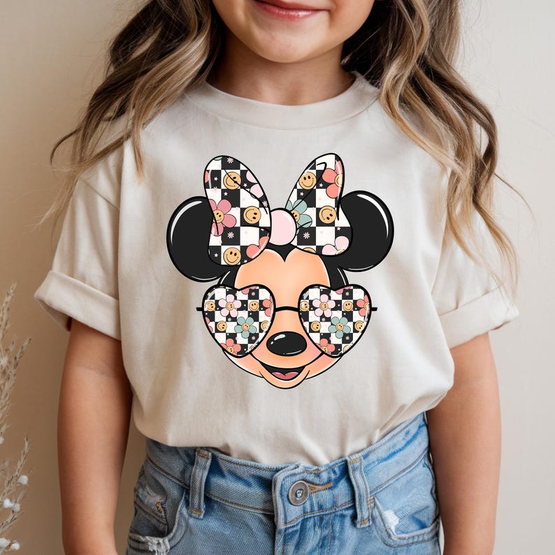 Puede incluir: Una camiseta de manga corta color crema con un gr&aacute;fico de una cabeza de rat&oacute;n de dibujos animados que lleva gafas en forma de coraz&oacute;n. El lazo y los cristales de las gafas tienen un patr&oacute;n de tablero de ajedrez blanco y negro, flores rosas y caras sonrientes amarillas.
