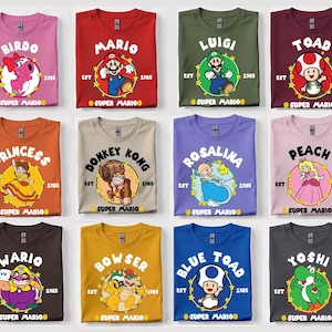 May include: Collection of Super Mario graphic t-shirts in various colors, each featuring a different character like Mario, Luigi, and Yoshi. The shirts have the character's name, the year 1985, and the words "Super Mario".