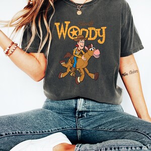 May include: Dark gray t-shirt featuring a graphic of Sheriff Woody riding a horse, with the text "Sheriff Woody" in orange and yellow. The shirt is a comfortable fit, perfect for fans of the animated movie.