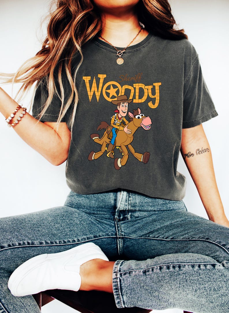 May include: Dark gray t-shirt featuring a graphic of Sheriff Woody riding a horse, with the text "Sheriff Woody" in orange and yellow. The shirt is a comfortable fit, perfect for fans of the animated movie.