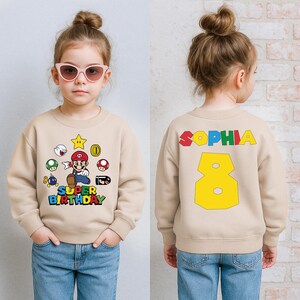 K&ouml;nnte beinhalten: Beiges Sweatshirt mit Super Mario-Motiv und dem Text "SUPER BIRTHDAY" auf der Vorderseite. Auf der R&uuml;ckseite stehen der Name "SOPHIA" und die Zahl "8" in bunten Farben.