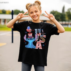 K&ouml;nnte beinhalten: Schwarzes T-Shirt mit einer blauen Cartoon-Figur, die einen patriotischen Hut tr&auml;gt, und dem Wort "USA" in Rot, Wei&szlig; und Blau. Das Design umfasst Sterne und Luftballons, die den Unabh&auml;ngigkeitstag feiern.