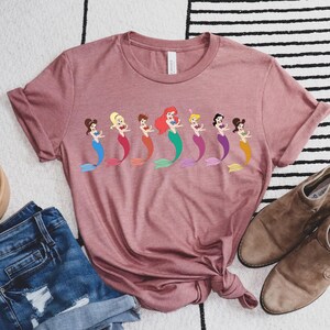 May include: A dusty rose t-shirt featuring a row of cartoon mermaids with colorful tails. The shirt is styled with denim shorts and brown ankle boots. A small potted succulent is also in the image.