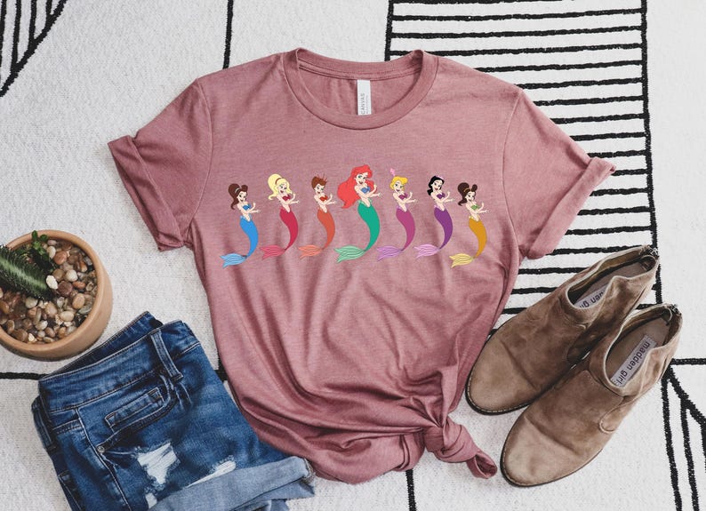 May include: A dusty rose t-shirt featuring a row of cartoon mermaids with colorful tails. The shirt is styled with denim shorts and brown ankle boots. A small potted succulent is also in the image.