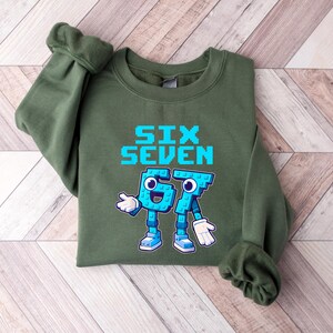 May include: An olive green sweatshirt with the words "SIX SEVEN" in pixelated blue letters. Below the text are two cartoon figures representing the numbers 6 and 7, in blue with white gloves and shoes. The sweatshirt has long sleeves.