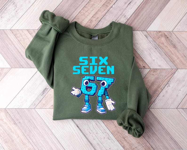 May include: An olive green sweatshirt with the words "SIX SEVEN" in pixelated blue letters. Below the text are two cartoon figures representing the numbers 6 and 7, in blue with white gloves and shoes. The sweatshirt has long sleeves.