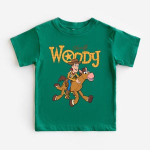 May include: A green t-shirt featuring a graphic of Sheriff Woody riding a horse, with the text "Sheriff Woody" in yellow. The design is centered on the front of the shirt.