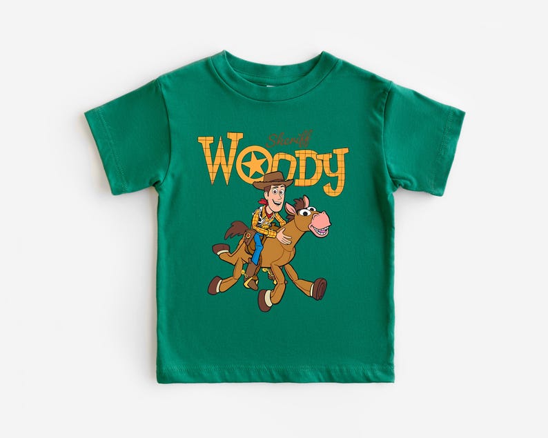 May include: A green t-shirt featuring a graphic of Sheriff Woody riding a horse, with the text "Sheriff Woody" in yellow. The design is centered on the front of the shirt.