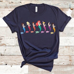 May include: Navy blue t-shirt featuring a row of cartoon mermaids with colorful tails. Each mermaid has a different hair color and tail color, including blue, red, green, purple, and orange. The shirt is made of soft material.