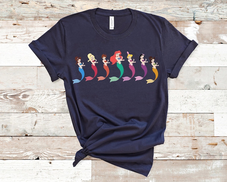 May include: Navy blue t-shirt featuring a row of cartoon mermaids with colorful tails. Each mermaid has a different hair color and tail color, including blue, red, green, purple, and orange. The shirt is made of soft material.