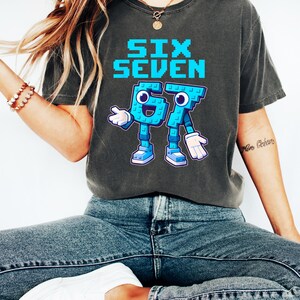 May include: Dark gray t-shirt with the words "SIX SEVEN" in pixelated blue letters. Below the text is a cartoon graphic of the numbers 6 and 7, also in blue, with arms, legs, and shoes. The shirt is worn with blue jeans and white sneakers.
