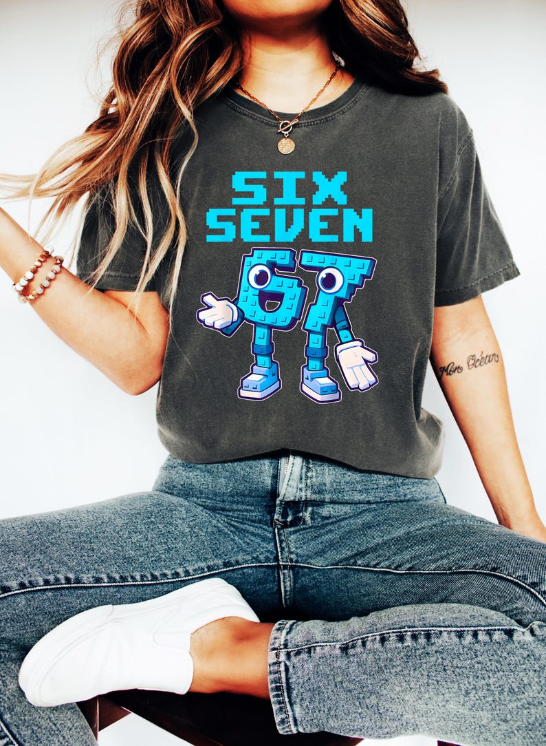 May include: Dark gray t-shirt with the words "SIX SEVEN" in pixelated blue letters. Below the text is a cartoon graphic of the numbers 6 and 7, also in blue, with arms, legs, and shoes. The shirt is worn with blue jeans and white sneakers.