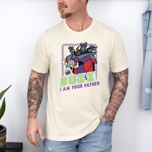 Puede incluir: Camiseta color crema con un gr&aacute;fico de Buzz Lightyear y Zurg con el texto "BUZZ I AM YOUR FATHER" en verde y blanco. El dise&ntilde;o est&aacute; enmarcado en un borde a cuadros morado y blanco.