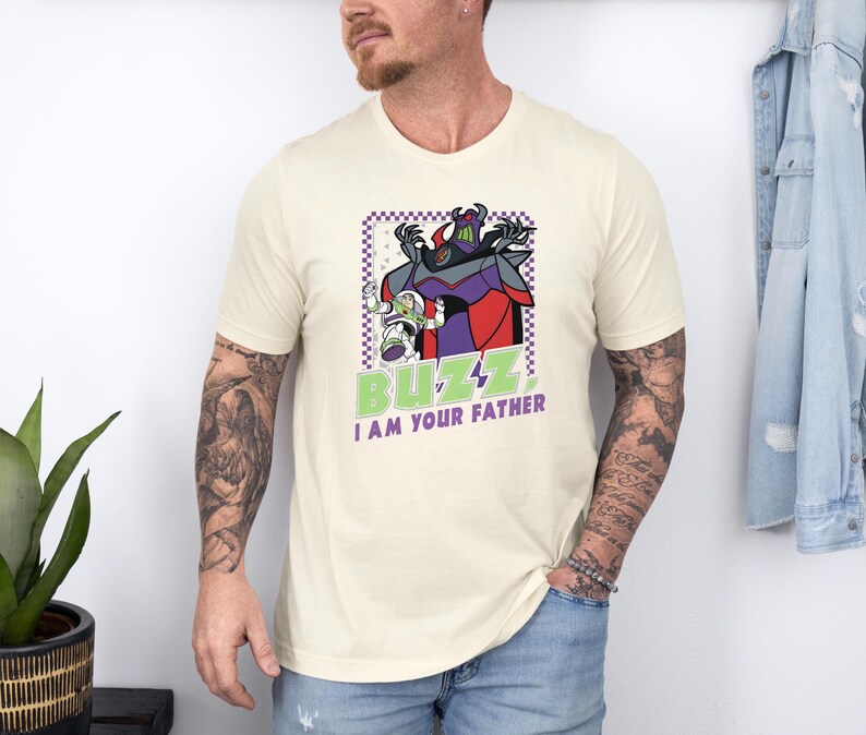 Puede incluir: Camiseta color crema con un gr&aacute;fico de Buzz Lightyear y Zurg con el texto "BUZZ I AM YOUR FATHER" en verde y blanco. El dise&ntilde;o est&aacute; enmarcado en un borde a cuadros morado y blanco.