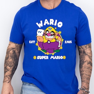 May include: Royal blue t-shirt featuring a cartoon image of Wario from Super Mario. The graphic includes Wario's image, the word "WARIO" above, and "EST 1985" and "SUPER MARIO" below. The shirt is a short-sleeved crew neck.