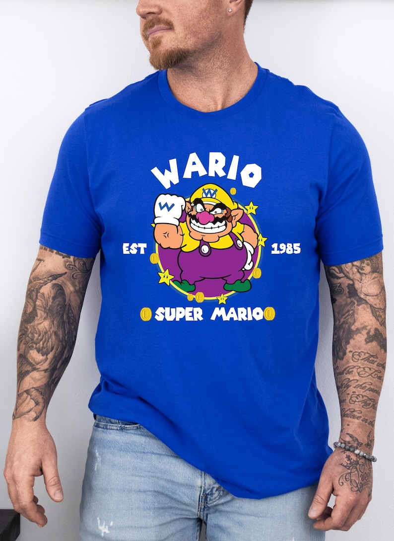 May include: Royal blue t-shirt featuring a cartoon image of Wario from Super Mario. The graphic includes Wario's image, the word "WARIO" above, and "EST 1985" and "SUPER MARIO" below. The shirt is a short-sleeved crew neck.