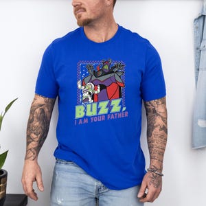 Puede incluir: Camiseta azul real con un gr&aacute;fico de Buzz Lightyear y Zurg de Toy Story, con el texto "Buzz, I am your father." El dise&ntilde;o est&aacute; enmarcado en un borde azul y verde. La camiseta tiene mangas cortas.