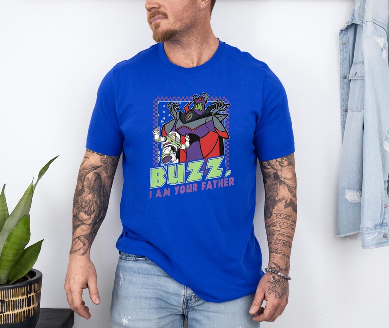 Puede incluir: Camiseta azul real con un gr&aacute;fico de Buzz Lightyear y Zurg de Toy Story, con el texto "Buzz, I am your father." El dise&ntilde;o est&aacute; enmarcado en un borde azul y verde. La camiseta tiene mangas cortas.