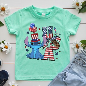 K&ouml;nnte beinhalten: Ein hellgr&uuml;nes T-Shirt mit einer blauen Cartoonfigur, die einen patriotischen Hut tr&auml;gt. Das Shirt hat einen "USA"-Schriftzug mit Sternen, Streifen und einem Leopardenmuster "S". Ballons und eine Rakete vervollst&auml;ndigen das Design.