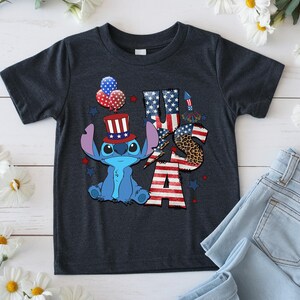 K&ouml;nnte beinhalten: Dunkelgraues T-Shirt mit einer blauen Cartoonfigur, die einen patriotischen Hut tr&auml;gt, und dem Text "USA" in Rot, Wei&szlig; und Blau. Das Design umfasst Ballons und Sterne, die den 4. Juli feiern.