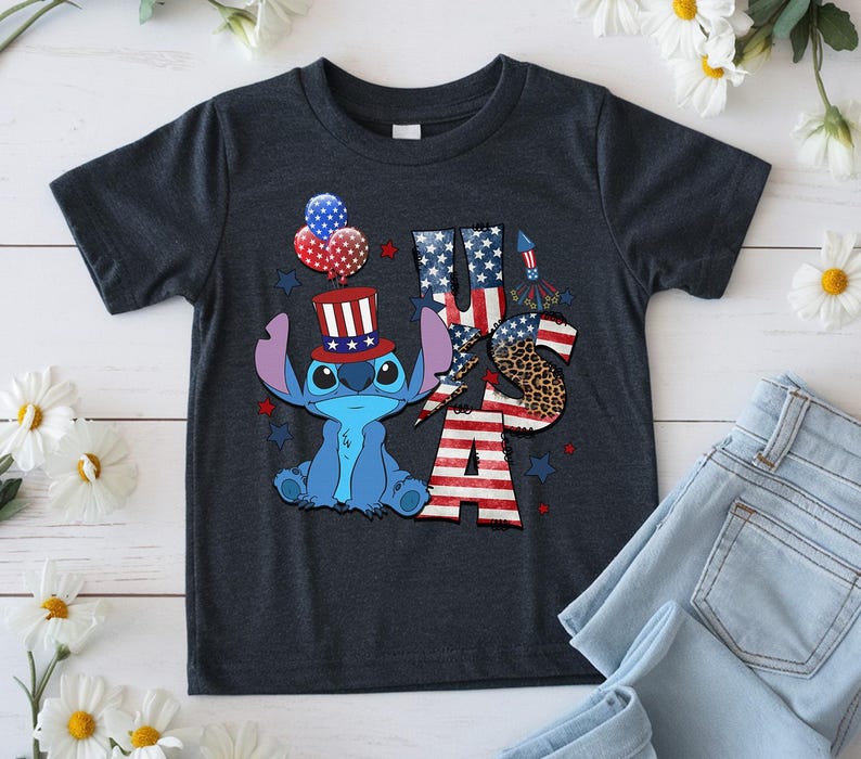 K&ouml;nnte beinhalten: Dunkelgraues T-Shirt mit einer blauen Cartoonfigur, die einen patriotischen Hut tr&auml;gt, und dem Text "USA" in Rot, Wei&szlig; und Blau. Das Design umfasst Ballons und Sterne, die den 4. Juli feiern.