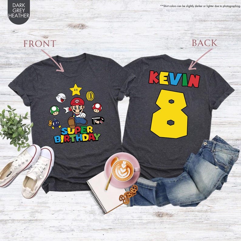 K&ouml;nnte beinhalten: Dunkelgraue Heather-T-Shirts mit Super Mario-Motiv. Die Vorderseite zeigt Mario und andere Charaktere mit dem Text "SUPER BIRTHDAY". Auf der R&uuml;ckseite stehen der Name "KEVIN" und die Zahl "8" in Gelb.