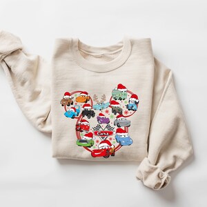 May include: Beige sweatshirt featuring a graphic of cartoon cars wearing Santa hats, arranged in a heart shape. The word "Cars" is in the center. The sweatshirt has long sleeves and a crew neck.