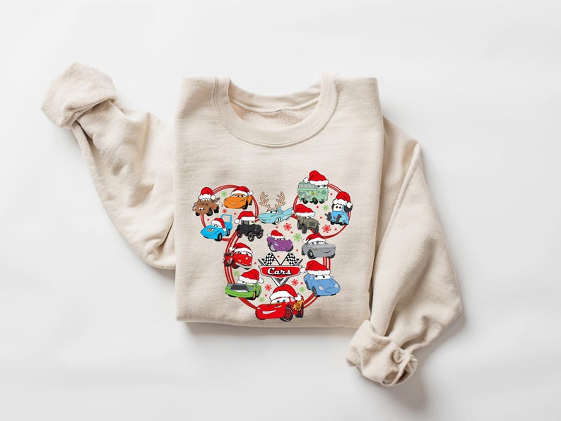 May include: Beige sweatshirt featuring a graphic of cartoon cars wearing Santa hats, arranged in a heart shape. The word "Cars" is in the center. The sweatshirt has long sleeves and a crew neck.