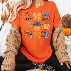 Puede incluir: Camiseta naranja con un dise&ntilde;o de Halloween que presenta personajes de dibujos animados, calabazas y hojas de oto&ntilde;o. La camiseta tiene cuello redondo y mangas cortas. El dise&ntilde;o incluye gr&aacute;ficos relacionados con Halloween en naranja, azul y marr&oacute;n.