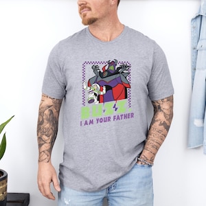 Puede incluir: Camiseta gris con un dise&ntilde;o de Buzz Lightyear y Zurg, con el texto "BUZZ v I AM YOUR FATHER". El dise&ntilde;o incluye morado, rojo y verde. La camiseta es de manga corta.