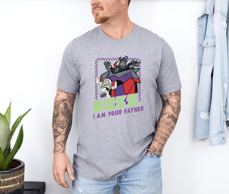 Puede incluir: Camiseta gris con un dise&ntilde;o de Buzz Lightyear y Zurg, con el texto "BUZZ v I AM YOUR FATHER". El dise&ntilde;o incluye morado, rojo y verde. La camiseta es de manga corta.