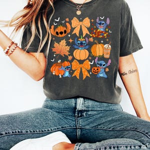 Puede incluir: Camiseta gris oscuro con un dise&ntilde;o de Halloween. El gr&aacute;fico incluye calabazas, hojas de oto&ntilde;o, lazos y un personaje de dibujos animados azul. La camiseta se usa con vaqueros azules y zapatillas blancas.