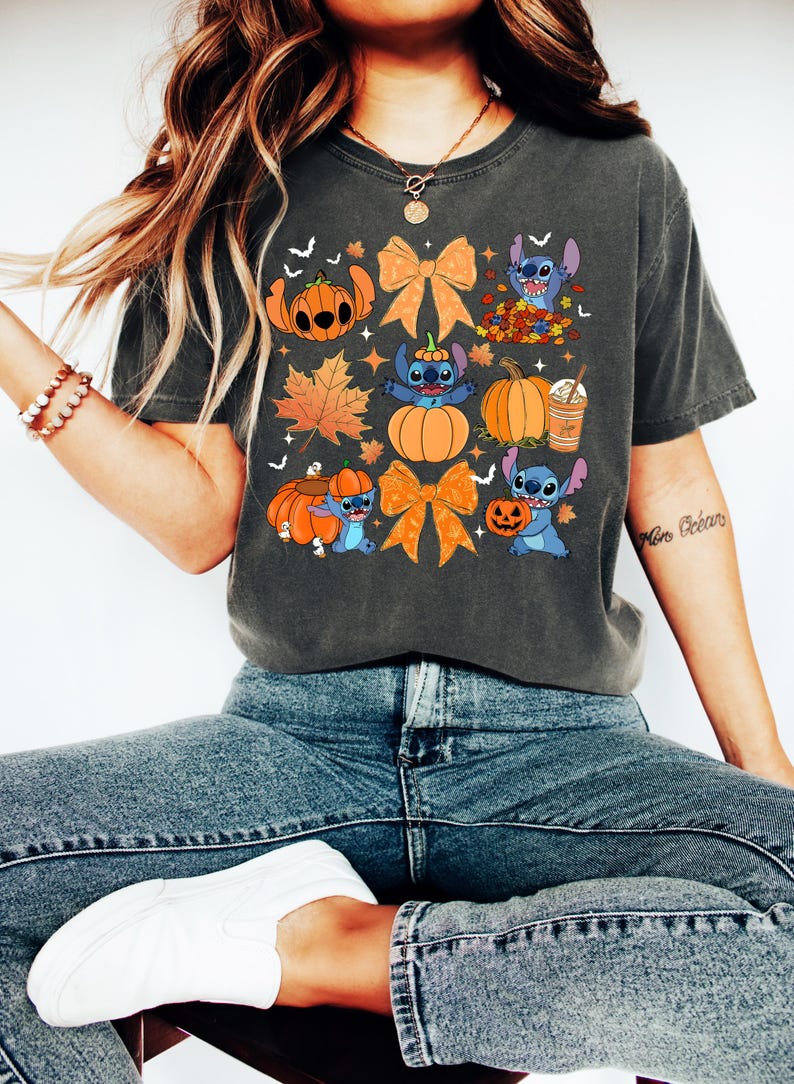 Puede incluir: Camiseta gris oscuro con un dise&ntilde;o de Halloween. El gr&aacute;fico incluye calabazas, hojas de oto&ntilde;o, lazos y un personaje de dibujos animados azul. La camiseta se usa con vaqueros azules y zapatillas blancas.