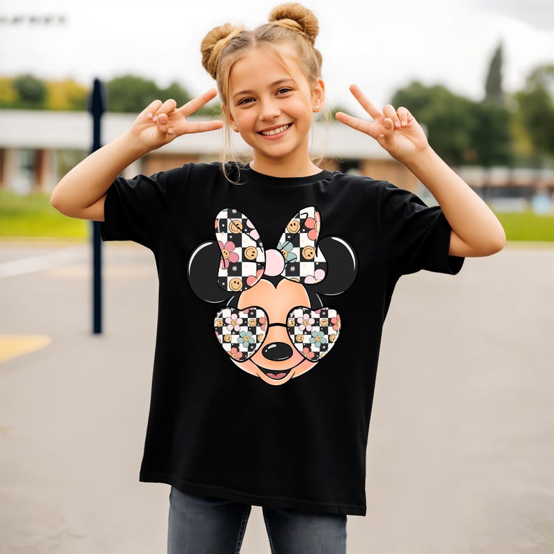 Puede incluir: Una camiseta negra con un gr&aacute;fico de una cabeza de rat&oacute;n de dibujos animados que lleva gafas en forma de coraz&oacute;n y un lazo. El dise&ntilde;o incorpora un patr&oacute;n de cuadros blancos y negros, peque&ntilde;as flores coloridas y caras sonrientes en el lazo y las gafas.