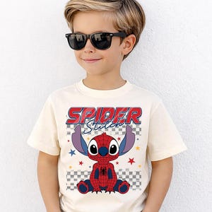May include: Cream-colored t-shirt featuring a graphic of Stitch as Spider-Man, with the text "SPIDER Stitch" in red and blue. The design includes a checkered background and star accents. The shirt is worn by a child wearing black sunglasses.