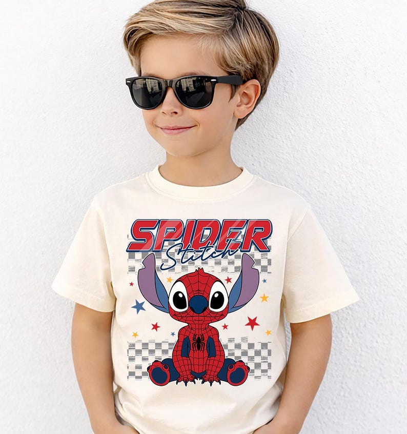 May include: Cream-colored t-shirt featuring a graphic of Stitch as Spider-Man, with the text "SPIDER Stitch" in red and blue. The design includes a checkered background and star accents. The shirt is worn by a child wearing black sunglasses.