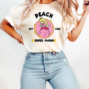 May include: Cream-colored t-shirt featuring a graphic of Princess Peach from Super Mario. The design includes the word "PEACH" above a circular image of the princess, with "EST 1985" and "SUPER MARIO" below. The princess is wearing a pink dress.