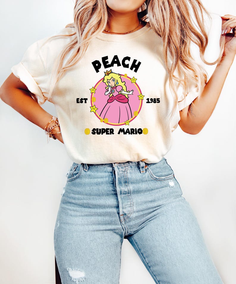 May include: Cream-colored t-shirt featuring a graphic of Princess Peach from Super Mario. The design includes the word "PEACH" above a circular image of the princess, with "EST 1985" and "SUPER MARIO" below. The princess is wearing a pink dress.