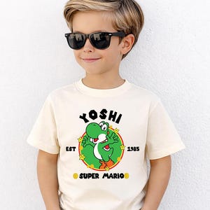 May include: Cream-colored t-shirt featuring a green Yoshi character graphic from Super Mario. The design includes the word "Yoshi" above the character, with "EST 1985" and "SUPER MARIO" below. The shirt is worn by a person wearing black sunglasses.