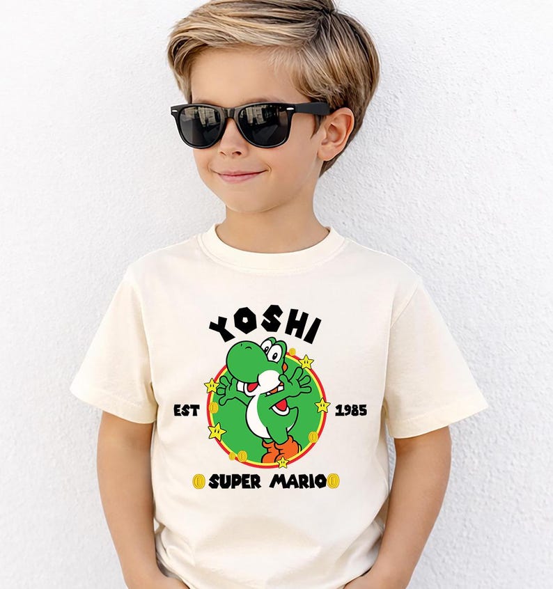 May include: Cream-colored t-shirt featuring a green Yoshi character graphic from Super Mario. The design includes the word "Yoshi" above the character, with "EST 1985" and "SUPER MARIO" below. The shirt is worn by a person wearing black sunglasses.