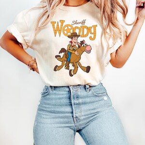 May include: Cream-colored t-shirt featuring a cartoon illustration of Sheriff Woody riding a horse. The text "Sheriff Woody" is printed above the image in a yellow, plaid-like font. The shirt is paired with light blue jeans.