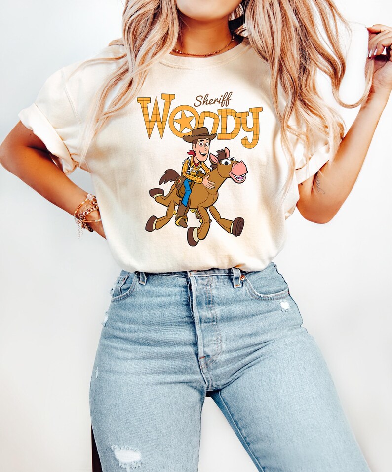 May include: Cream-colored t-shirt featuring a cartoon illustration of Sheriff Woody riding a horse. The text "Sheriff Woody" is printed above the image in a yellow, plaid-like font. The shirt is paired with light blue jeans.